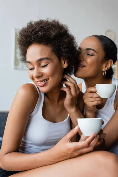 Счастливая американская пара лесбиянок, держащая чашки кофе, улыбаясь дома — стоковое фото