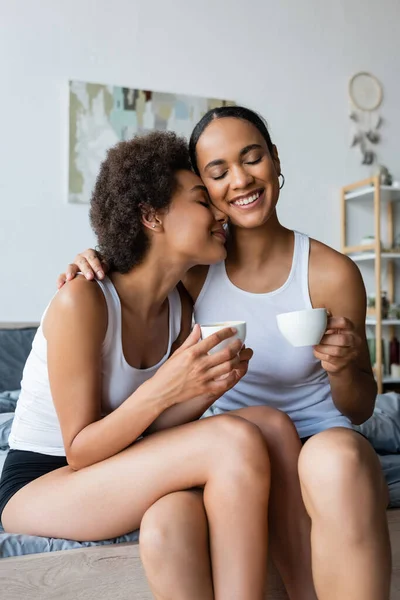 Alegre pareja afroamericana lesbiana sosteniendo tazas de café mientras sonríe en casa - foto de stock