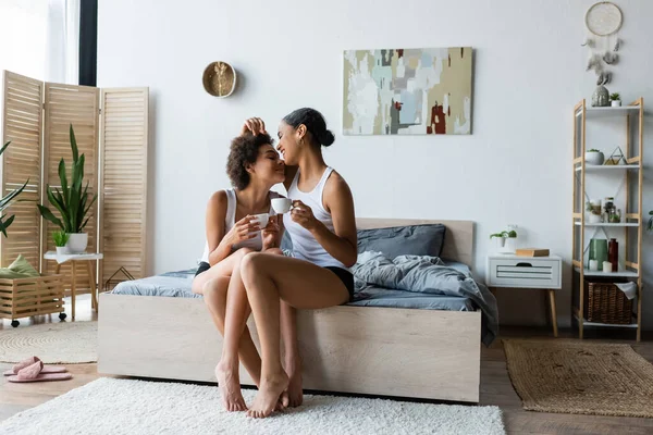 Full length of cheerful lesbian african american couple holding cups of coffee while sitting on bed — Stock Photo