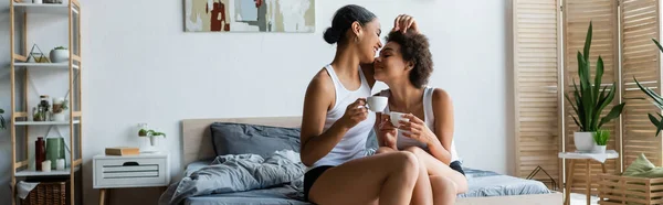 Alegre pareja afroamericana lesbiana sosteniendo tazas de café mientras está sentado en la cama, pancarta - foto de stock