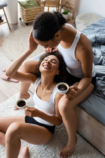 Vista superior de mulheres americanas africanas lésbicas alegres abraçando e segurando xícaras de café no quarto — Fotografia de Stock