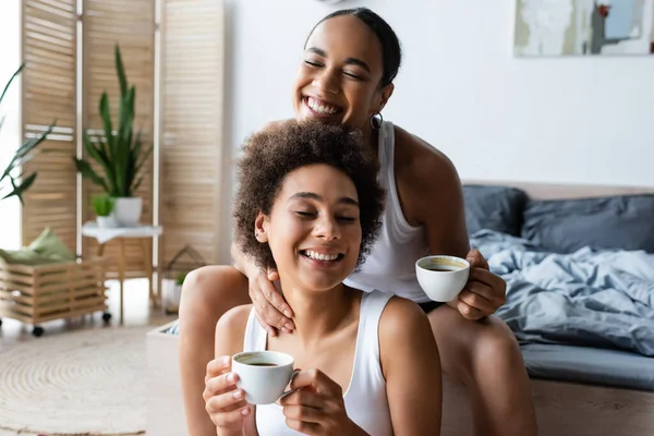 Fröhliche lesbische afrikanisch-amerikanische Frau umarmt Freundin mit Tasse Kaffee — Stockfoto