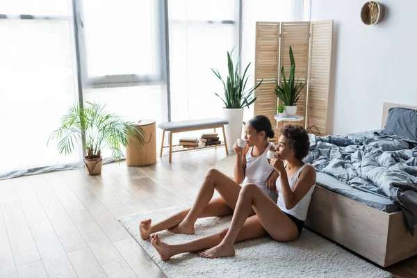 Longitud completa de jóvenes lesbianas africano americano pareja beber café mientras sentado cerca de la cama - foto de stock