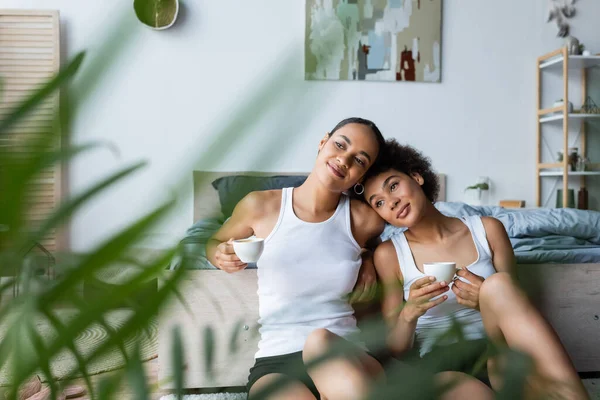 Pleased lesbian african american women holding cups of coffee while sitting near plant on blurred foreground — Stock Photo