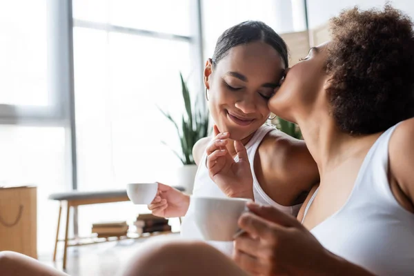 Lesbiana africana americana mujer besos mejilla de sonriente novia con taza de café - foto de stock