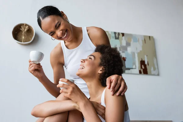 Happy african american lesbian woman smiling and hugging girlfriend with cup of coffee — Stock Photo