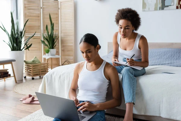 Young african american woman with notebook looking at laptop near girlfriend in bedroom — Stock Photo