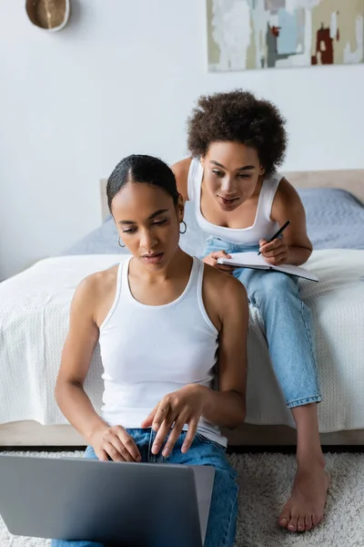 Кудрявая африканская американка с блокнотом, смотрящая на ноутбук рядом с подругой в спальне — стоковое фото
