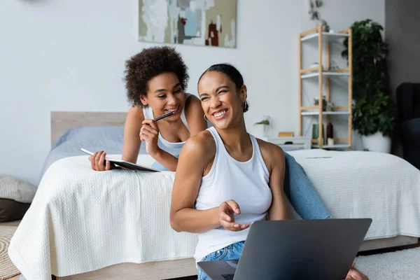 Glückliche afrikanisch-amerikanische Frau mit Notizbuch lächelnd in der Nähe ihrer Freundin, die fern zu Hause arbeitet — Stockfoto