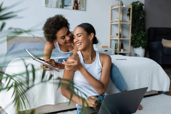 Heureux afro-américaine femme assise avec ordinateur portable et pointant avec la main à portable près de petite amie — Photo de stock