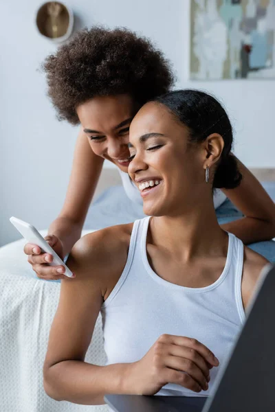 Mujer afroamericana feliz sosteniendo teléfono inteligente cerca de novia positiva con el ordenador portátil - foto de stock