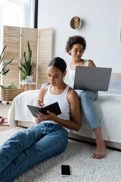 Afro-américaine femme écrit dans un cahier près de petite amie de travail de la maison avec ordinateur portable — Photo de stock