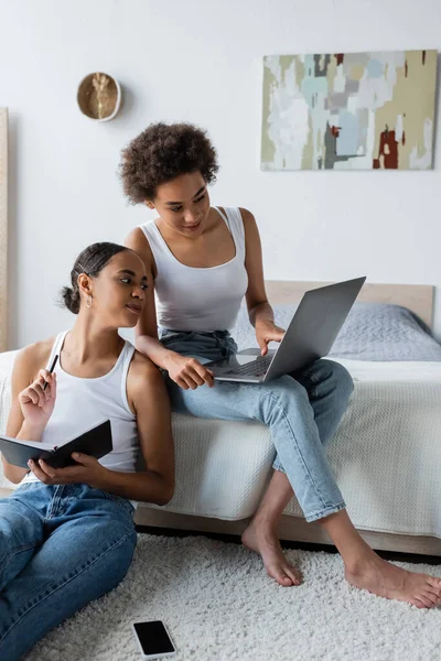Afro-américaine femme tenant un cahier et regardant ordinateur portable près de petite amie bouclée travaillant à la maison — Photo de stock