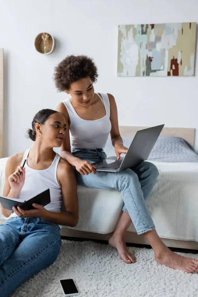 Mujer afroamericana sosteniendo portátil y mirando a la computadora portátil cerca de novia rizada trabajando desde casa - foto de stock