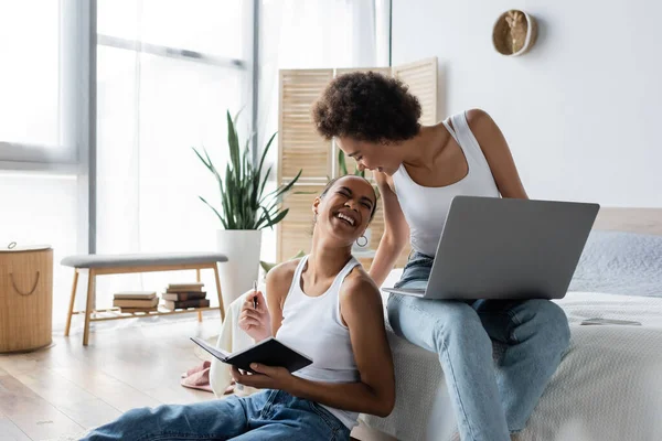 Glückliche afrikanisch-amerikanische Frau mit Laptop schaut fröhliche Freundin mit Notizbuch an — Stockfoto