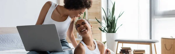 Happy african american woman with laptop looking at cheerful girlfriend laughing in bedroom, banner — Stock Photo