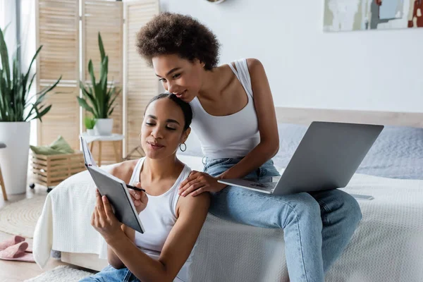 Happy african american woman with laptop looking at notebook in hands of cheerful girlfriend — Stock Photo