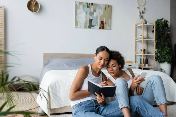 Lesbian african american woman holding smartphone and looking at notebook in hands of girlfriend — Stock Photo