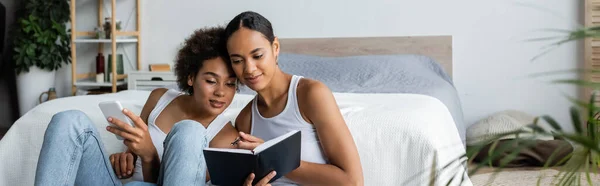 Mujer afroamericana lesbiana sosteniendo teléfono inteligente y mirando el cuaderno en manos de su novia, pancarta - foto de stock