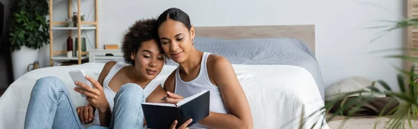 Lesbienne afro-américaine femme tenant smartphone et regardant cahier dans les mains de petite amie, bannière — Photo de stock