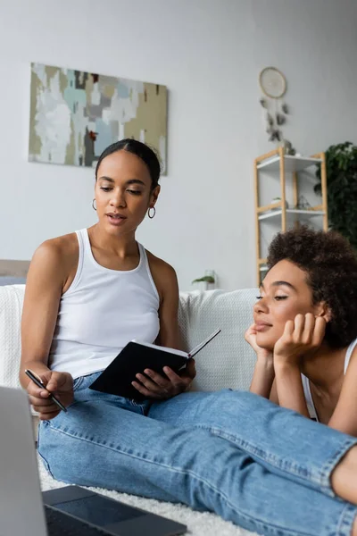 Lesbian african american woman holding notebook while talking with girlfriend and looking at laptop — Stock Photo