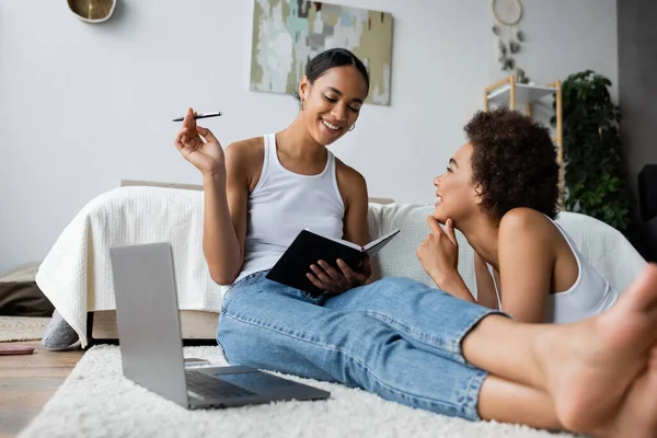 Felice e lesbica donna afro-americana guardando la ragazza vicino al computer portatile in camera da letto — Foto stock