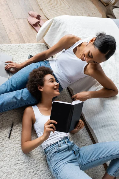 Top view of curly african american woman lying on laps of cheerful girlfriend and holding notebook — Stock Photo