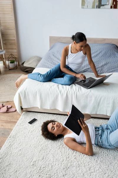 Curly african american woman lying on carpet and looking at notebook near girlfriend using laptop on bed — Stock Photo