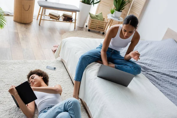 Joven afroamericana mujer acostada en la alfombra y mirando el cuaderno cerca de novia usando el ordenador portátil en la cama - foto de stock