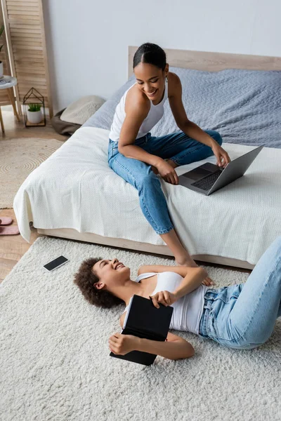 Happy african american woman lying on carpet and looking girlfriend using laptop on bed — Stock Photo