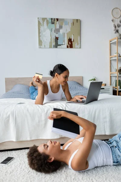 Joven afroamericana mujer acostada en la alfombra cerca de novia de compras en línea con el ordenador portátil y tarjeta de crédito - foto de stock