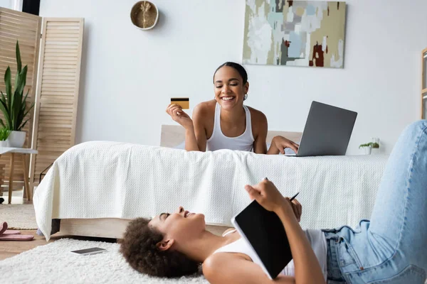 Jeune femme afro-américaine couchée sur le tapis et regardant la petite amie heureuse tenant la carte de crédit tout en utilisant un ordinateur portable — Photo de stock