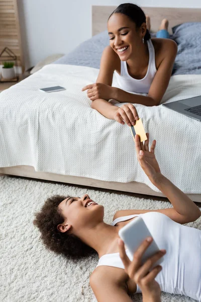 Junge Afroamerikanerin liegt auf Teppich und gibt Freundin Kreditkarte — Stockfoto