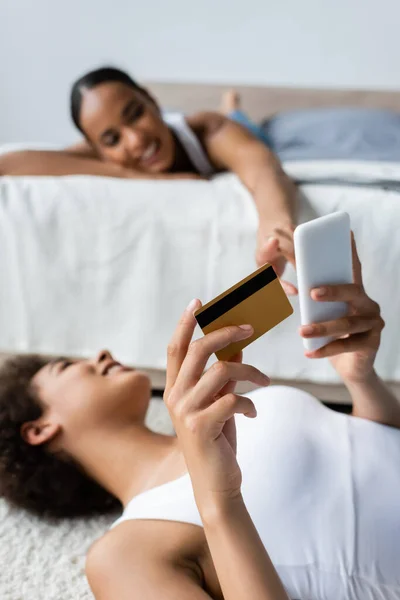 Jeune femme afro-américaine en utilisant un smartphone tout en tenant la carte de crédit et en regardant la petite amie sur fond flou — Photo de stock