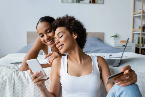 Heureuse africaine américaine lesbienne femme en utilisant smartphone tout en tenant carte de crédit près de petite amie — Photo de stock