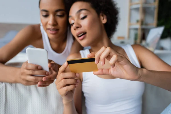 Stupéfait afro-américain lesbienne femme tenant carte de crédit près de petite amie avec smartphone — Photo de stock
