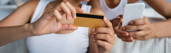 Обрезанный вид на африканскую американскую лесбиянку, держащую кредитку рядом с подругой со смартфоном — стоковое фото