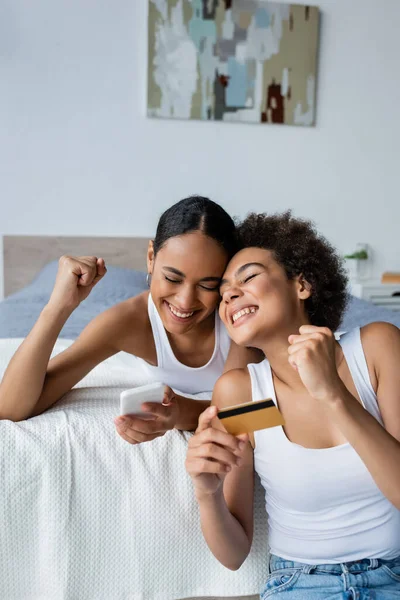 Счастливая американская лесбиянка из Африки, держащая кредитку рядом с взволнованной подругой со смартфоном — стоковое фото