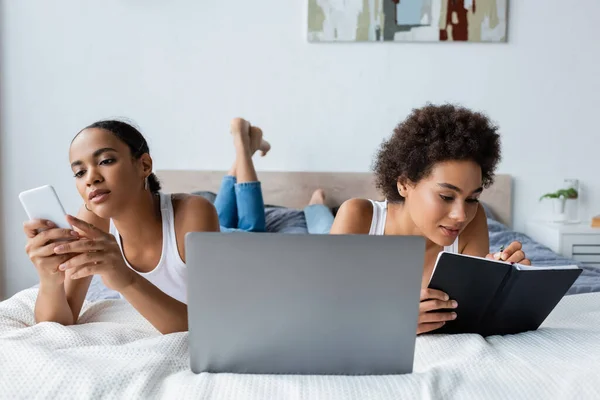 African american lesbian woman using smartphone while girlfriend writing in notebook near laptop on bed — Stock Photo
