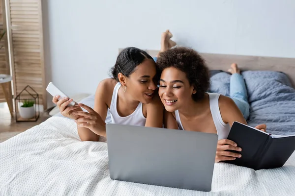 Mujer lesbiana afroamericana feliz sosteniendo teléfono inteligente mientras habla con su novia cerca del ordenador portátil en la cama - foto de stock