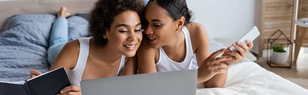 Happy african american lesbian woman holding smartphone while talking with girlfriend near laptop, banner — Stock Photo
