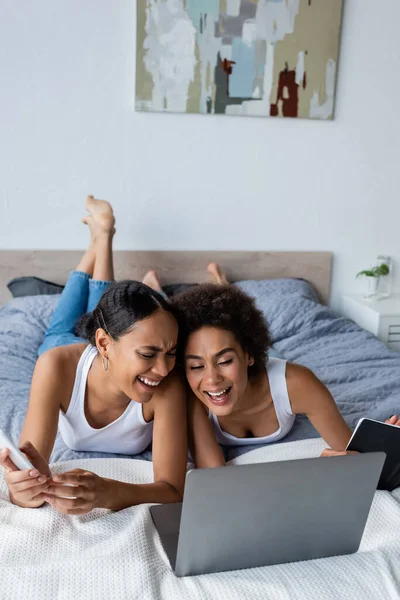 Feliz africano americano lesbiana pareja riendo cerca laptop en cama - foto de stock