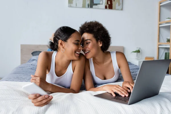 Feliz africano americano lesbiana pareja sonriendo cerca laptop en cama - foto de stock