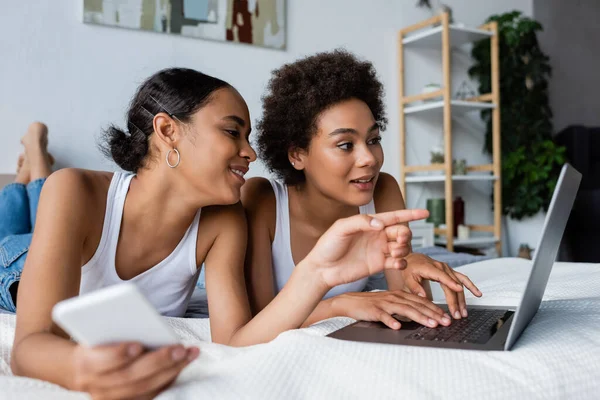 Happy african american lesbian woman holding smartphone while pointing at laptop near girlfriend — Stock Photo