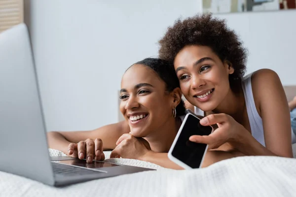 Feliz africano americano lesbiana mujer celebración smartphone con pantalla en blanco mientras busca portátil cerca de novia - foto de stock