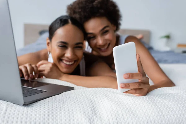 Sonriente afroamericana lesbiana mujer mostrando smartphone a novia cerca de portátil en la cama - foto de stock