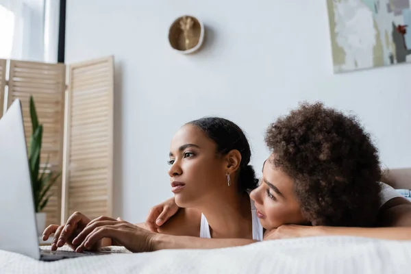 Curiosa africana americana lesbiana mujer mirando novia usando laptop en cama - foto de stock