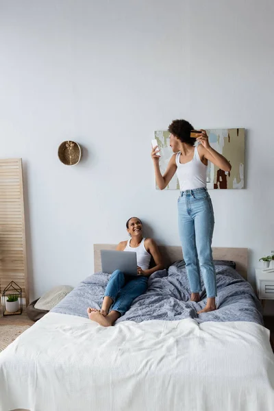 Возбужденная американская лесбиянка, держащая смартфон и кредитную карту, стоя на кровати рядом с подругой — стоковое фото