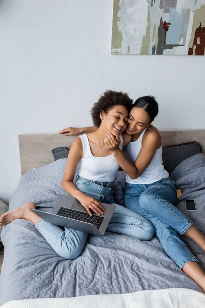 Американская лесбийская пара из Африки обнимается возле ноутбука и смартфона на кровати — стоковое фото