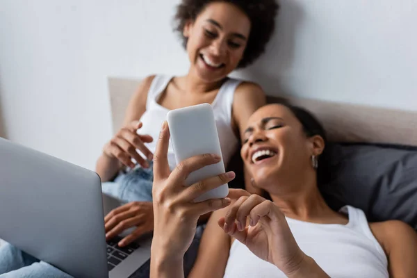 Positive african american lesbian woman holding smartphone girlfriend in bedroom — Stock Photo